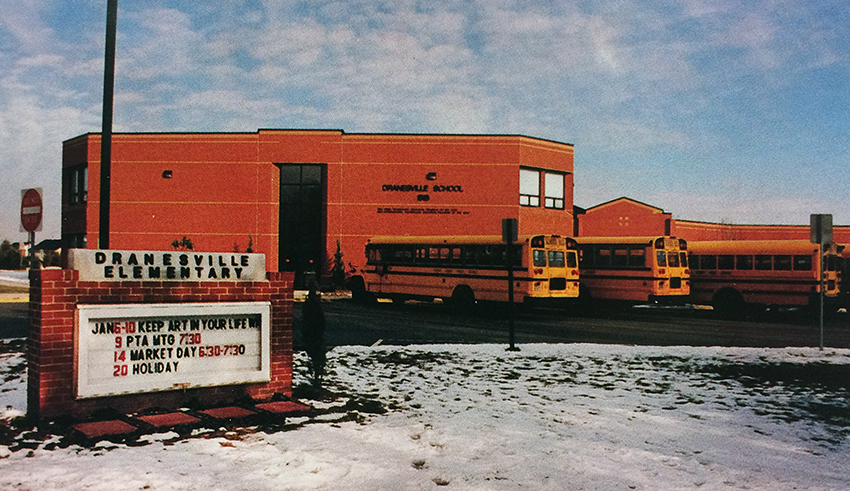 : Photograph of the exterior of Dranesville Elementary from the 1996 to 1997 yearbook. Several school buses are parked in front of the building and there is a blanket of snow on the ground.