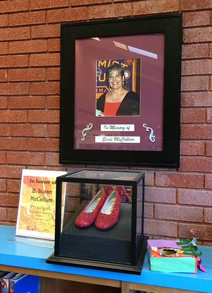 Photograph of the memorial to Principal McCallum in Dranesville’s library. There is a portrait of Ms. McCallum on the wall and beneath her are ruby slippers in a glass case.